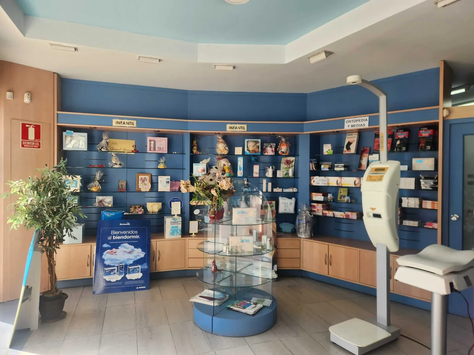Interior de la farmacia de Raquel Barcos en Zizur Mayor: estanterías azules con productos infantiles y de ortopedia y medias, vitrina redonda de cristal y báscula con tallímetro